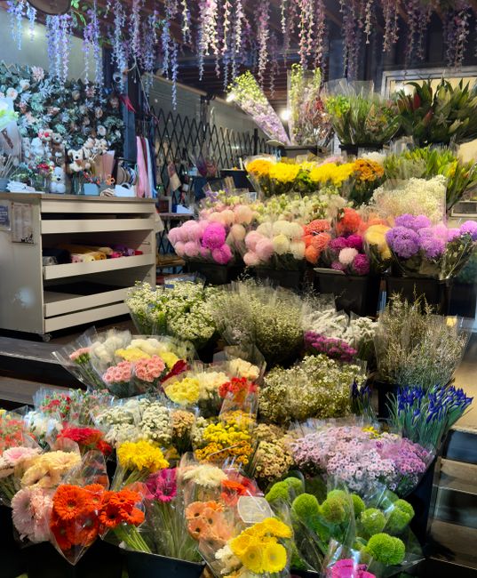 A vibrant display of fresh flowers in a florist shop, featuring a variety of blooms including pink, white, yellow, purple, and orange chrysanthemums, roses, and lilies, all arranged in clear wrapping 