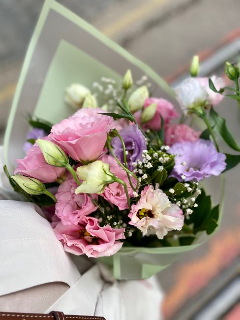 A hand holding a bouquet of fresh flowers featuring pink lisianthus, white roses, and light purple lisianthus, accented with small white baby's breath flowers and green foliage. The bouquet is wrapped