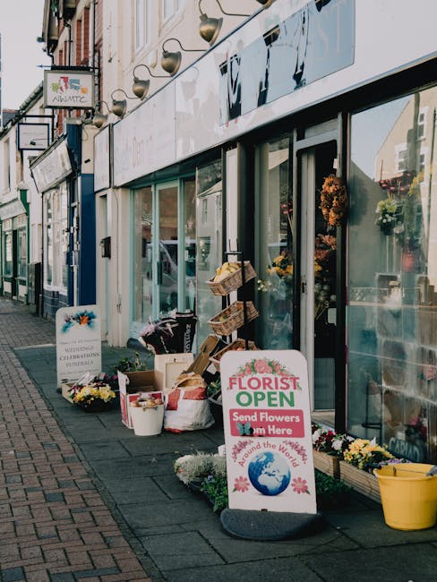 The storefront of floristnoakhill.co.uk displaying an array of floral arrangements and supplies outside the shop. The scene includes a variety of fresh flowers in white buckets, including chrysanthemu
