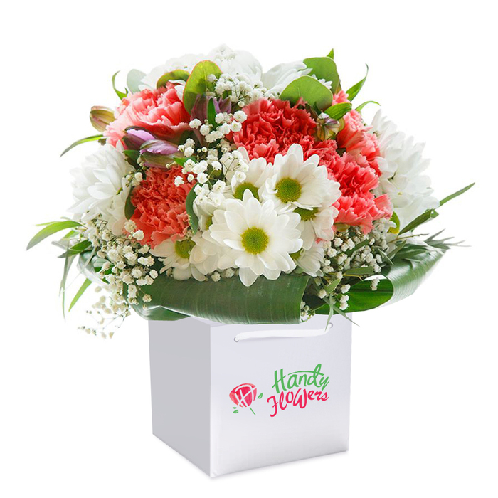 Perfectly arranged within a crisp white, square-shaped gift bag, this vibrant bouquet brings together the loveliest colours and textures-a true reflection of Noak Hill's inviting character. Stately coral and flaming red carnations lead the ensemble, their abundant, ruffled petals offering a rich, tactile elegance. Throughout, white daisies with sunny yellow-green centers provide a delightful pop, evoking the gentle charm of Noak Hill's village gardens. Wisps of white gypsophila thread softly through the bouquet, imparting a wispy delicacy that lightens the mood. Sprigs of purple-tinged buds draw the eye, adding depth and subtle interest, as if glancing into hidden corners of the countryside. All is embraced by abundant deep green foliage, each leaf glossy and broad, encircling the flowers like protective arms. The bouquet's rounded shape and lively mix radiate freshness-its artistry presented upright in a white bag with strong ribbon handles, set against a clear, bright white background. Ideal for special moments-birthdays, anniversaries, or simply to show you care-this arrangement by Florist Noak Hill offers an elegant gift infused with local pride, artistry, and the promise of a smile at first glance.