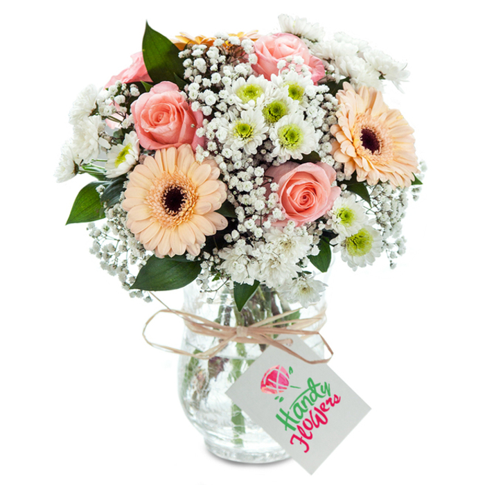 Set against a luminous white backdrop that brings out every detail, this lavish bouquet is a tribute to Noak Hill's soft, rural elegance. A careful blend of classic, peach-tinted roses-each one full and beautifully open-anchors the heart of the arrangement, their abundant petals hinting at romance and enduring grace. Clusters of cheerful peach-orange gerbera daisies stand prominently, their wide, sun-kissed petals and deep brown centers emanating a sense of belonging and local cheer, much like the vibrant community spirit felt on Noak Hill's high streets. Between these star blooms, fluffy nodules of white chrysanthemum infuse the bouquet with subtle texture, their tight green-yellow centers peeking through, while a generous scattering of fine, white baby's breath lends a light, airy volume reminiscent of morning fog rolling across local fields. Sparse, dark green eucalyptus foliage weaves through the composition, adding scent and a tactile freshness that echoes the nearby woodlands. The flowers are arranged in a crystal-clear vase with a gentle, bulbous base narrowing elegantly at the top, exposing the green stems as they rest in pristine water. A slim wrap of raffia-tinted twine is knotted around the vase's neck, and from it dangles a bright, rectangular white gift tag-an inviting blank canvas for any heartfelt message. Professionally crafted with care, this bouquet captures the tranquil beauty and heartfelt warmth of Noak Hill, perfect for marking special occasions with a touch of local artistry and timeless style.