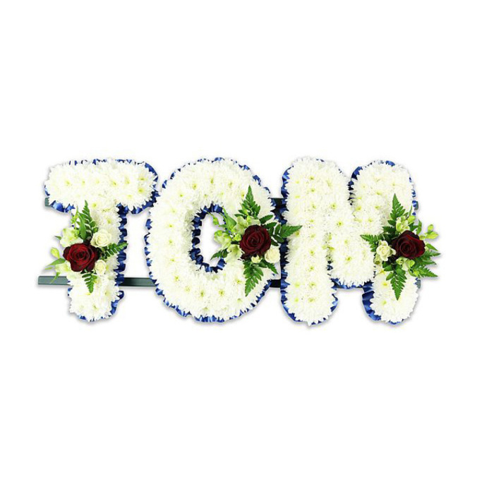 This personalised funeral tribute spells TOM in bold, softly rounded letters made from a profusion of small white chrysanthemums, the blooms packed densely to produce a pillowy, tactile surface that looks as if you could rest a hand upon it. Around the perimeter of each letter a royal blue ribbon has been applied with precise, even tension, forming a crisp frame that accentuates the letters' contours and lends a composed, formal air. Each letter carries a modest, symmetrical spray: centrally placed dark red roses serve as poignant focal points, their velvety petals suggesting deep affection and mourning, while accompanying ivory spray roses and feathery fern fronds soften the composition and add layers of subtle green. The understructure - a thin dark metal bar - is discreetly visible just beneath the base, indicating the letters are meant to be displayed upright on an easel or at a graveside. Photographed against a pure white background with soft directional light, the arrangement's textures and tonal contrasts are clear: the white chrysanthemums read pristine; the royal blue ribbon holds visual weight; the red roses draw the eye and the greenery creates gentle movement. The overall effect is ceremonial yet intimate, a crafted expression of remembrance suited to services across Noak Hill and neighbouring Havering communities. There is an implied hush in the composition, the quiet scent of chrysanthemums and the gentle perfume of roses forming an unseen comfort for those who come to honour a life.