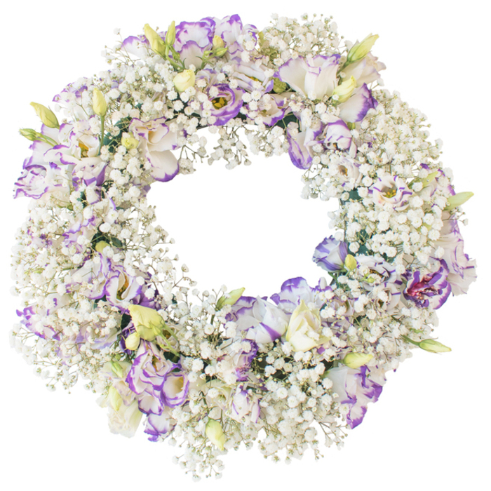 This image shows a classic circular funeral wreath lying against a pristine white backdrop, every element of its construction clearly visible. The wreath has been fully covered with stems of white baby's breath, so densely packed that the underlying base disappears beneath a halo of tiny blossoms. The baby's breath creates an intricate, lace-like surface that feels soft and airy, like a ring of fine foam or a collar of snow. Set into this cloud of white are generously spaced lisianthus blooms, chosen in a palette of white touched with purple. Each lisianthus opens in overlapping, ruffled petals, the outer edges brushed with tones of lavender and violet that gently deepen as they curve outward, giving the flowers a painterly quality. A number of blooms are at their peak, revealing layered centres with a hint of depth and shadow, while several buds and partially open flowers add movement and a sense of natural progression. The wreath forms a smooth, unbroken ring with a clear central opening, a shape that naturally suggests cycles of life and the enduring bond between the living and those they have lost. The lighting is bright and even, casting no distractions on the crisp background, so the viewer's focus remains on the interplay between the fine texture of the baby's breath and the bolder forms of the lisianthus. This refined, tranquil arrangement feels suited to remembrance ceremonies across Noak Hill, whether placed near the altar of a village church or resting by a graveside, offering a quiet, graceful symbol of sympathy and respect.